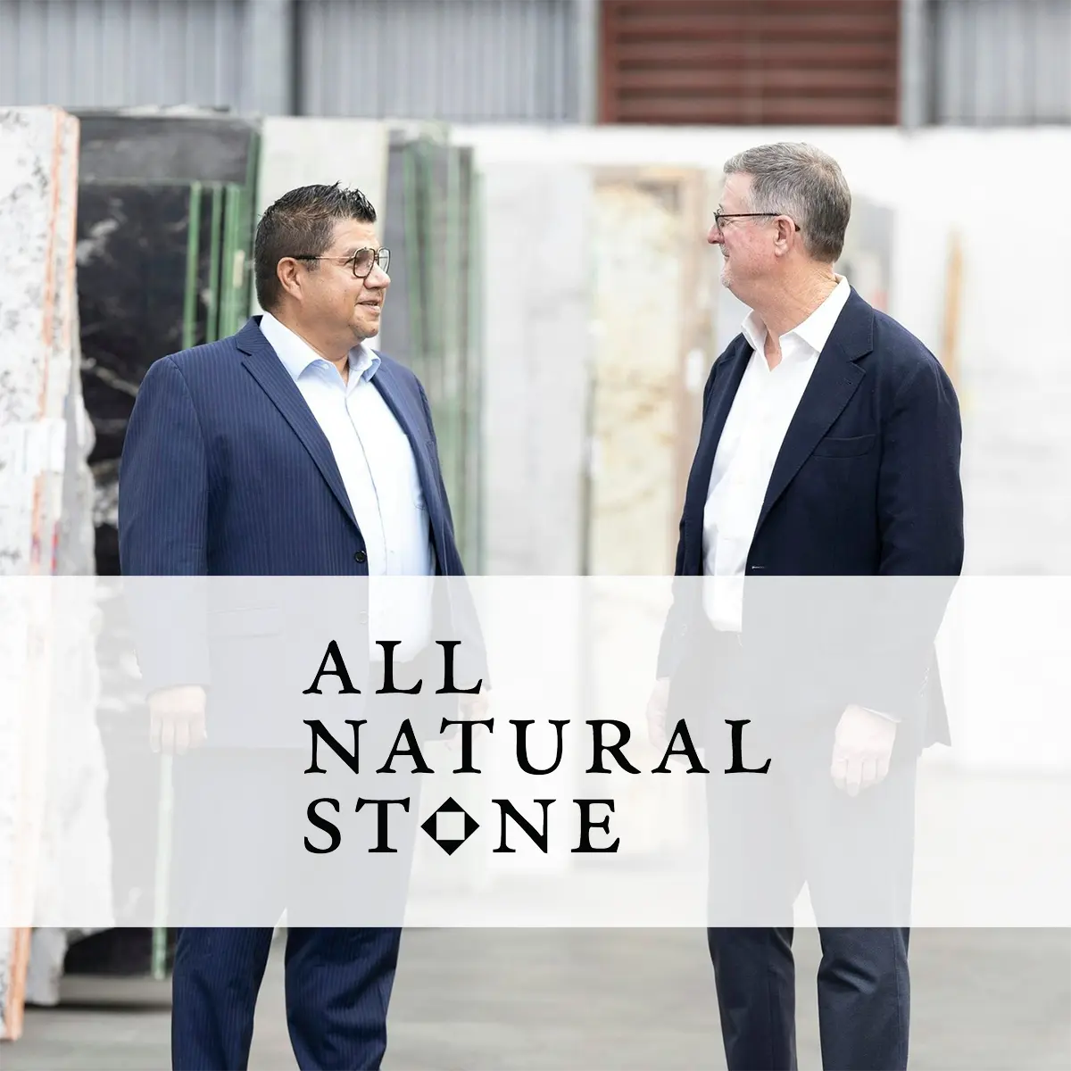 Two All Natural Stone team members in suits smile while talking inside a stone slab showroom, surrounded by large marble and natural stone slabs.